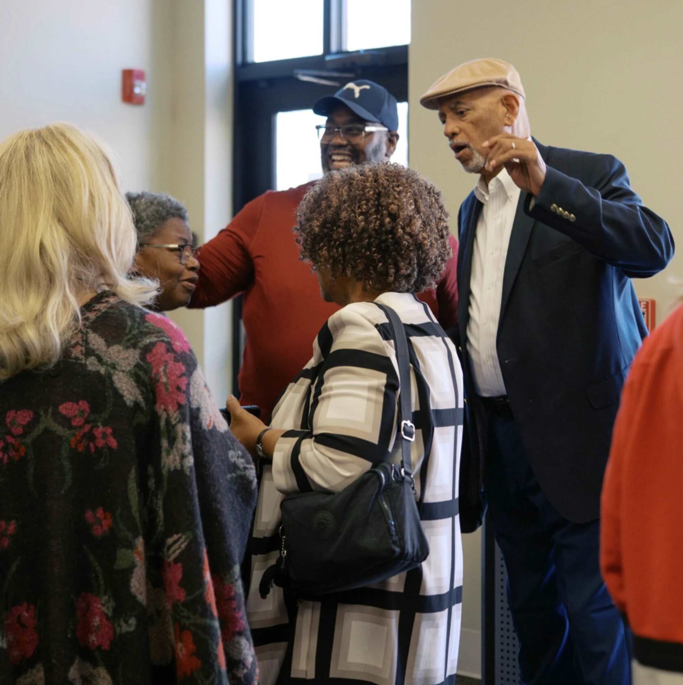 A group of older adults, mostly Black, stand together talking indoors. A man in a cap gestures as he speaks, while others listen and smile.